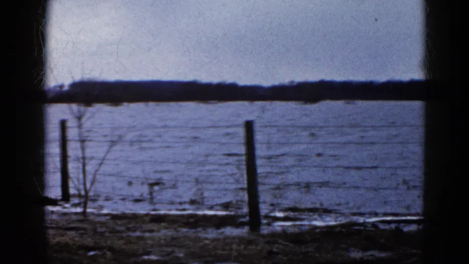 1961 Wauconda, Illinois view across an icy lake from behind a dark frame, with barbed wire, fence posts, sparse brush, and distant trees.