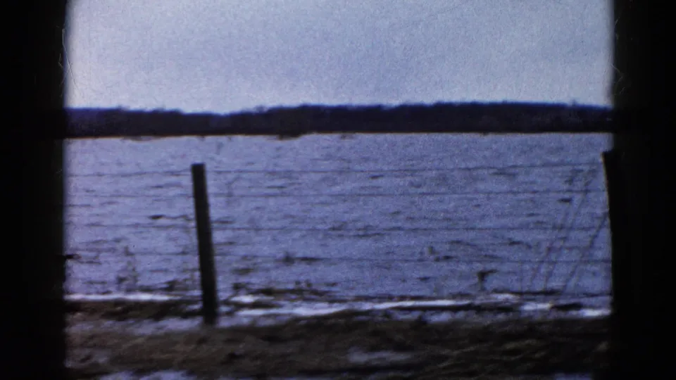 1961 Wauconda, Illinois frame of a frozen lake shoreline through dark framing, with a leaning fence post, barbed wire, snow traces, and brush.