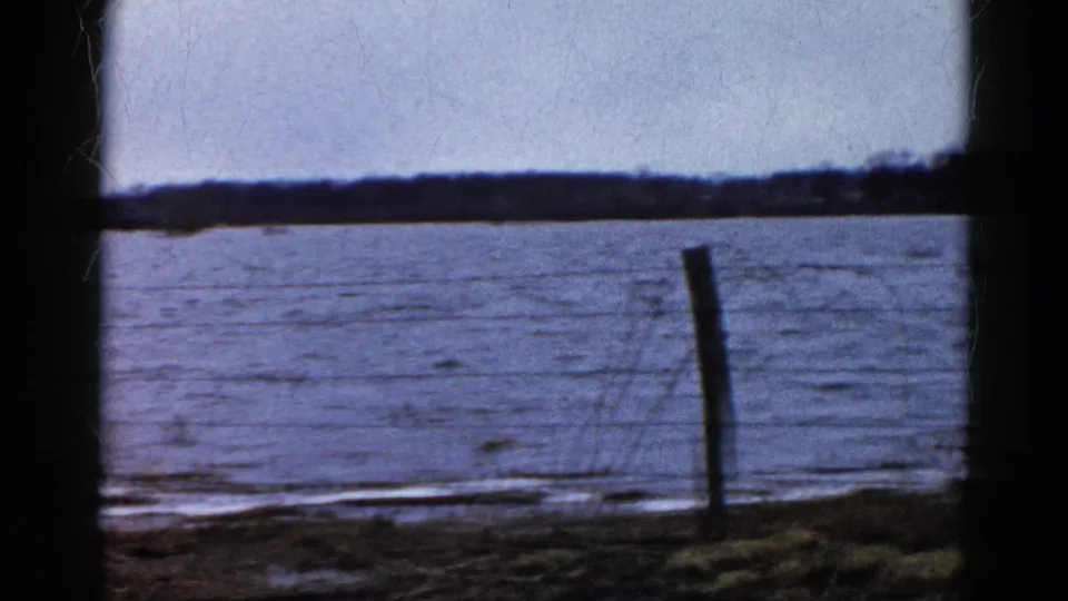 1961 Wauconda, Illinois home-movie frame showing a frozen lake beyond barbed wire and a fence post, with sparse shoreline brush and dark framing.