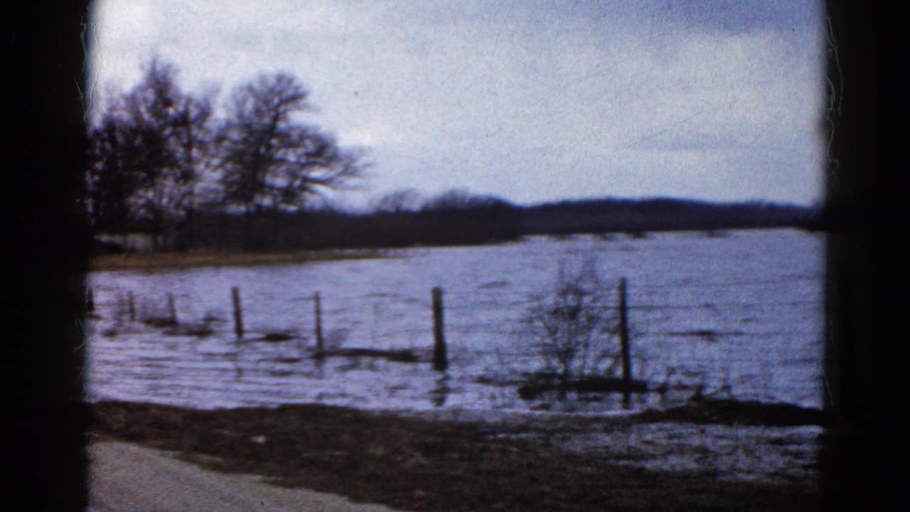 Floodwater crosses a rural fence line beside a road under an overcast sky.