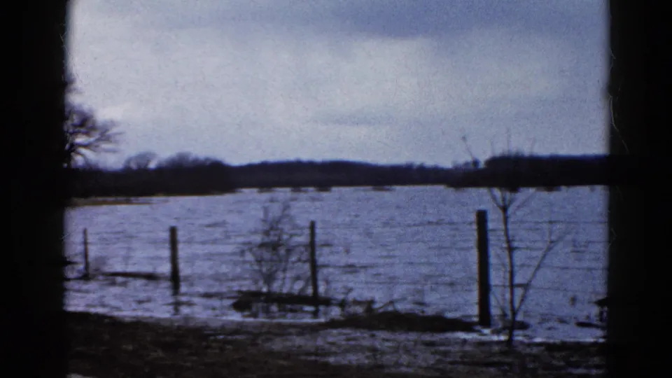 Flooded rural ground in Wauconda, Illinois, with water passing through fence posts and brush beneath a gray 1961 sky.
