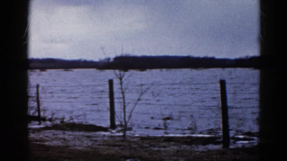 Rural Wauconda flood scene from 1961 showing water behind a fence line, bare shrubs, distant trees, and muted 8mm color.