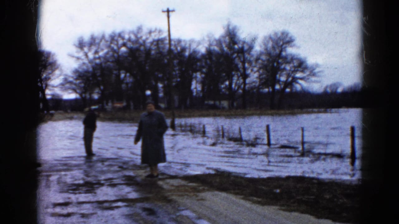 Two adults face high water beside fence posts and bare trees.