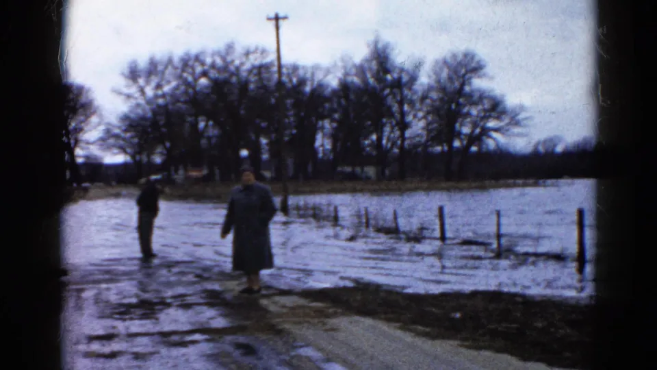 Two adults in heavy coats stand beside rising water on a rural Wauconda roadside, with fence posts partly submerged.