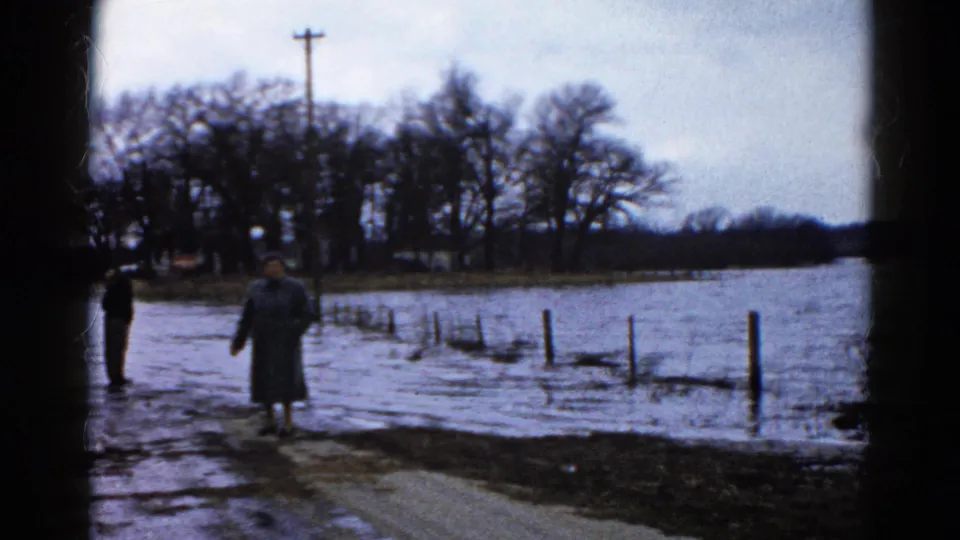 A coat-wearing adult stands by a flooded rural road in the supplied Wauconda, Illinois 1961 home-movie scene.