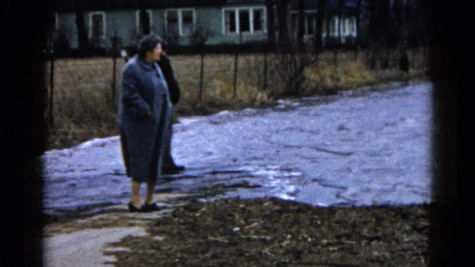 In Wauconda, Illinois, 1961, a woman in a long coat stands near a wet roadside as a house and bare trees sit in the background.