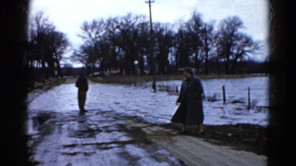 The 1961 Wauconda home movie shows a man and woman separated by a flooded road, with bare trees, fence posts, and standing water nearby.