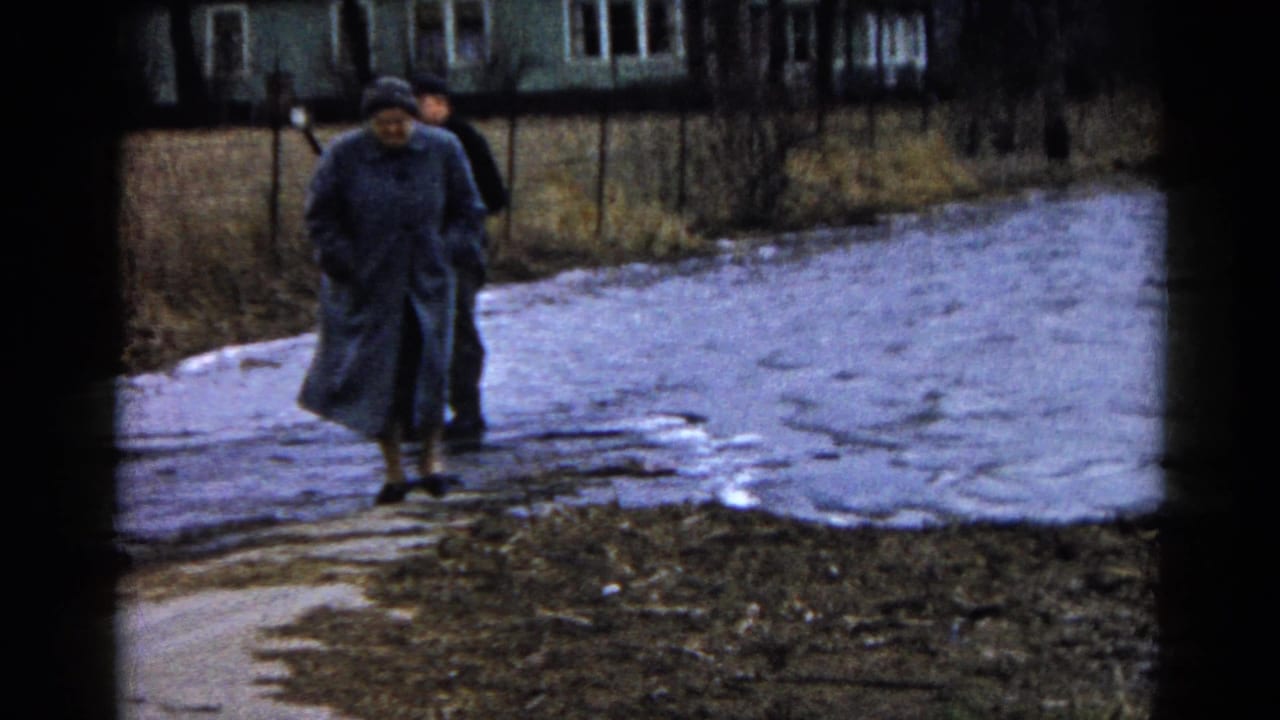 Two adults in coats walk near a muddy path beside water.
