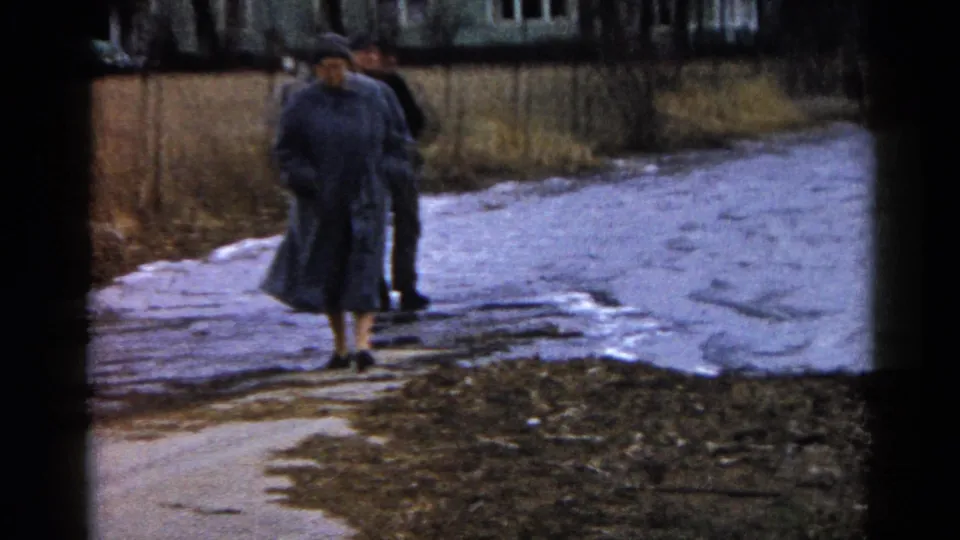 A woman in a long gray coat walks toward the camera while a second adult in dark clothing follows near the muddy water edge in 1961 Wauconda.