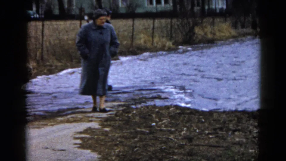 A woman in a long gray coat and hat walks beside the water in Wauconda, Illinois, with muddy ground, snow patches, and a fence behind her.