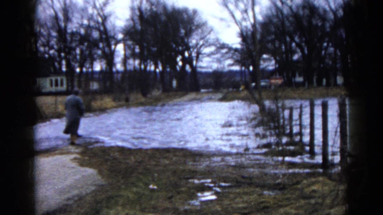 Adult in a coat walking near pooled meltwater on a muddy winter path.