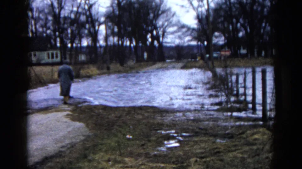The adult pauses near standing water on a muddy path in 1961 Wauconda, with bare trees, fence posts, and low houses behind.