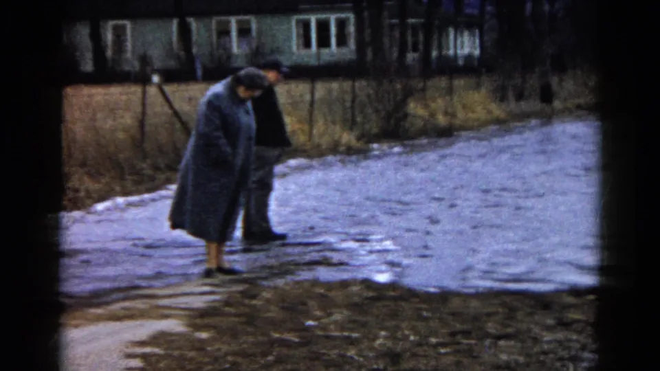 Two adults in dark winter clothing stand near the flooded path in Wauconda, Illinois, with one leaning down to inspect the wet ground.