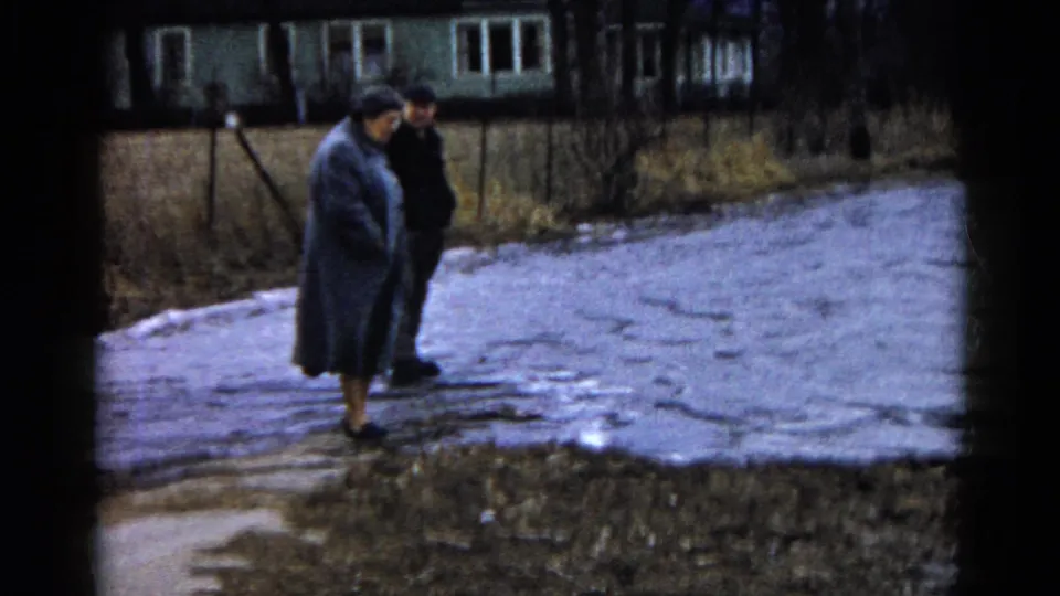 Two adults stand together beside pooled meltwater on a muddy Wauconda path, with houses, bare trees, and winter grass in the background.