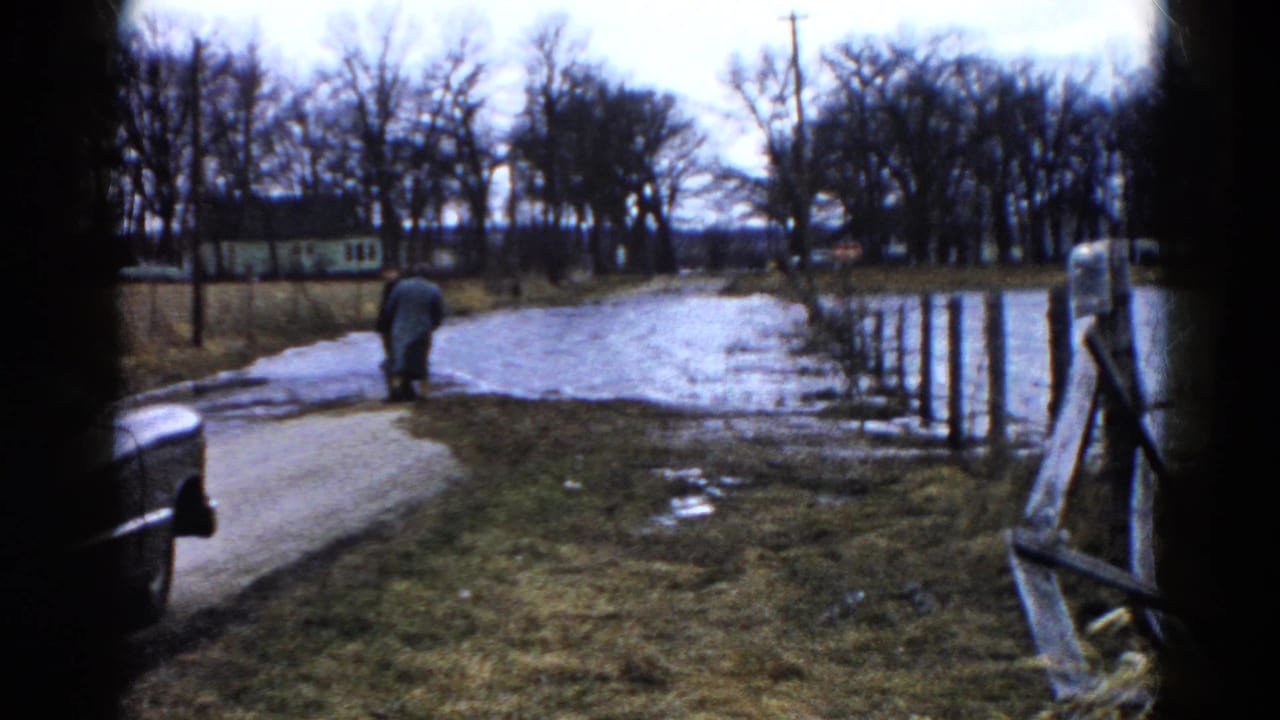 Two adults walk away beside a flooded road near a parked car and bare trees.