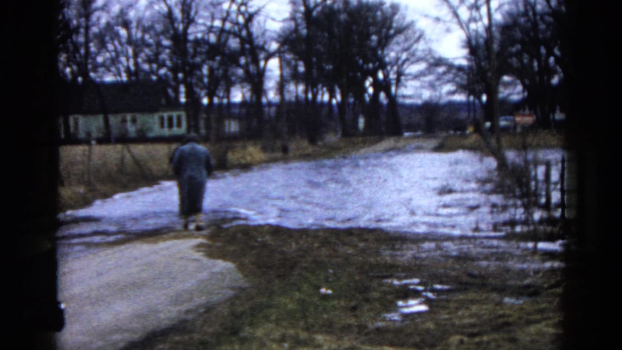 A distant adult in a long coat walks along a wet road toward floodwater in Wauconda, Illinois, in 1961.