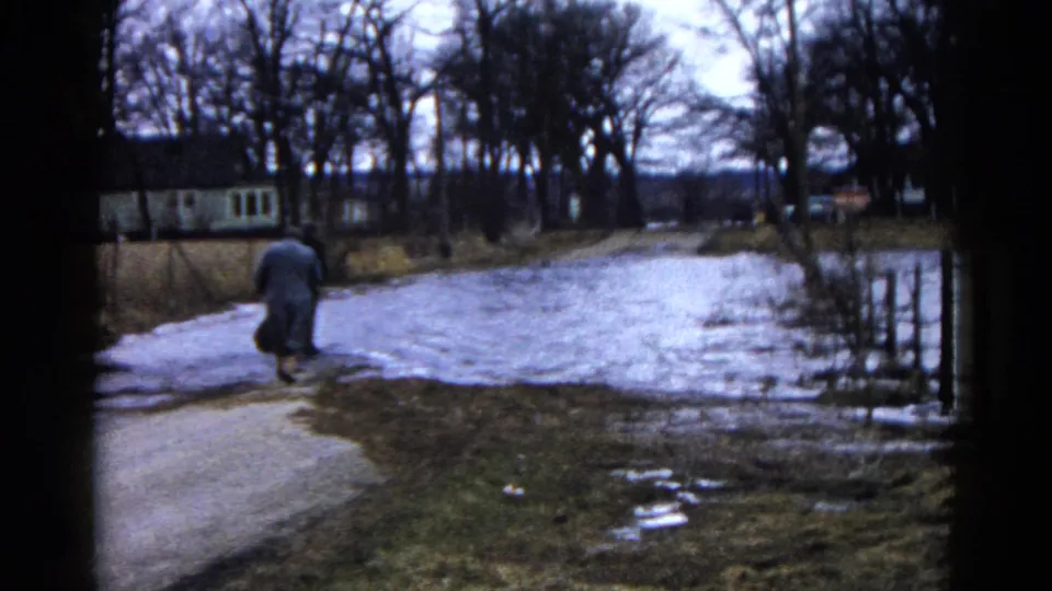 An adult walks beside floodwater on a rural Wauconda road, with bare trees and distant houses visible in the 1961 home movie frame.