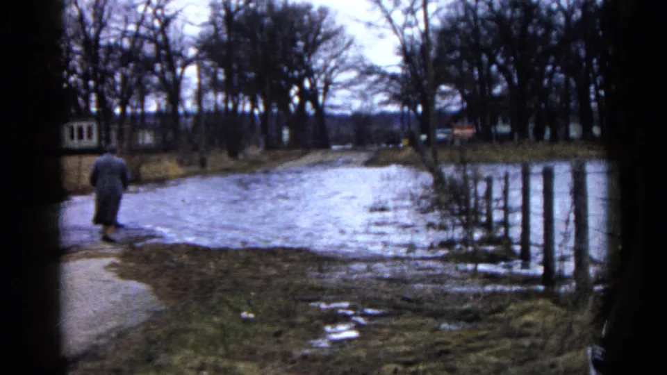 The 1961 Wauconda home-movie view shows a distant adult near water covering the road, with fence posts and leafless trees around the scene.