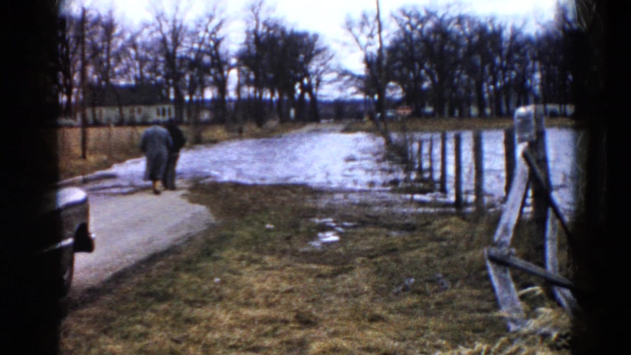Person in a long coat walking away on a muddy roadside beside standing water.