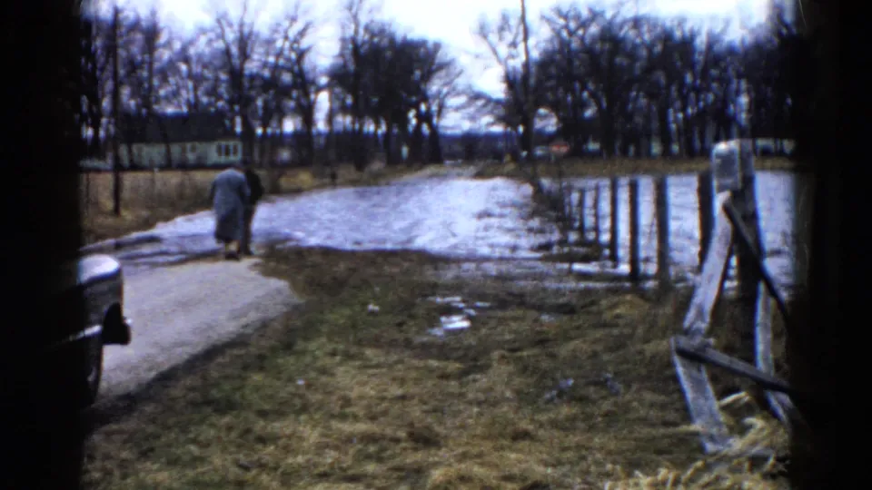 The person continues away from the camera on a muddy Wauconda roadside, with standing water, fence rails, bare trees, and grass visible in the frame.