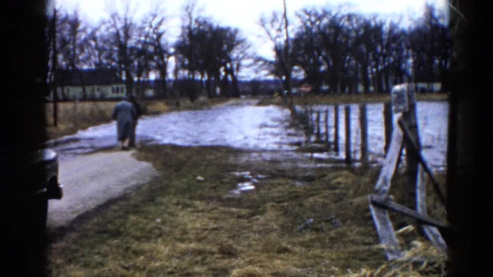 A 1961 home-movie frame shows the walker farther down the muddy road near icy water, with a wooden fence, bare trees, and rural grassland around them.