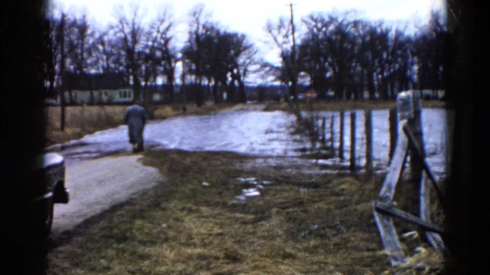 At the end of the clip, the person is farther away on the muddy Wauconda road, with standing water, fence posts, bare trees, and grass still visible.