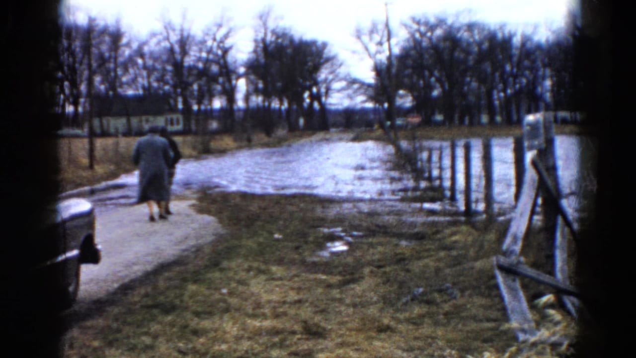 Two adults walk away on a wet cement walkway beside high water and a wooden fence.