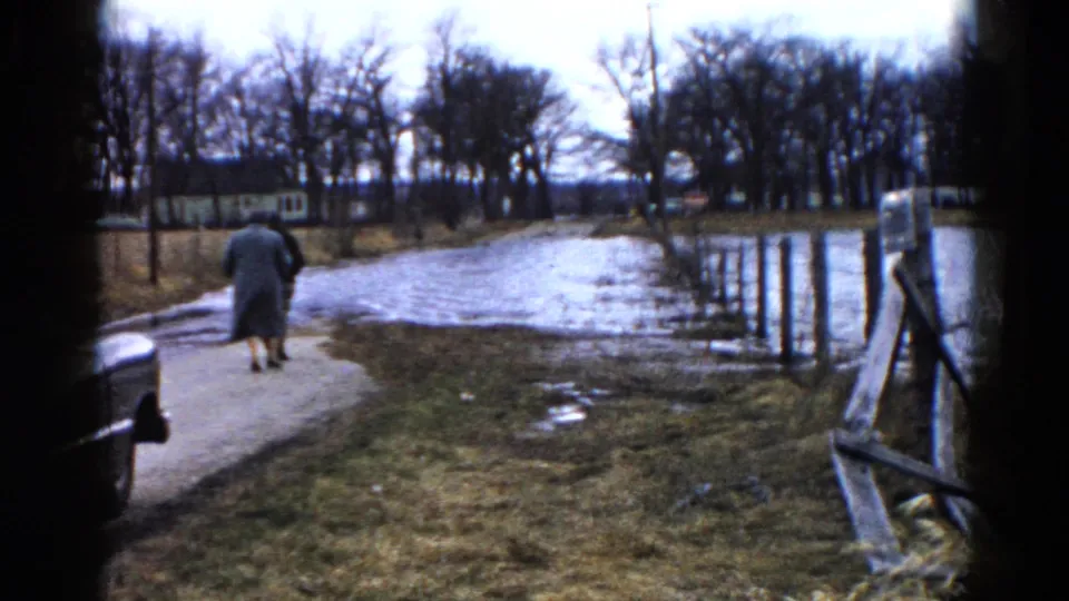 In 1961 Wauconda, Illinois, two adults in coats walk away along a wet path near water, bare trees, and a wooden fence.
