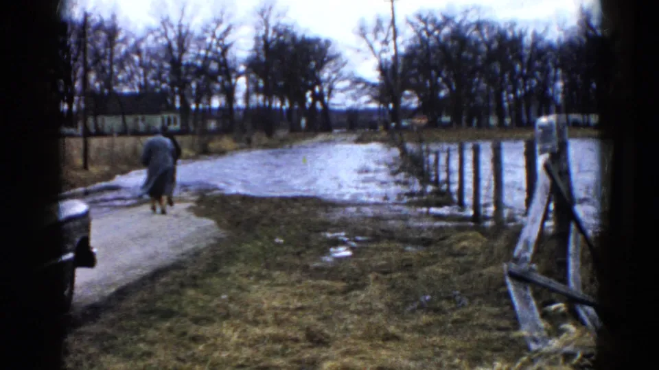 Two adults in long coats walk farther away in the 1961 Wauconda scene, near water, grass, bare trees, and a partial car at frame edge.
