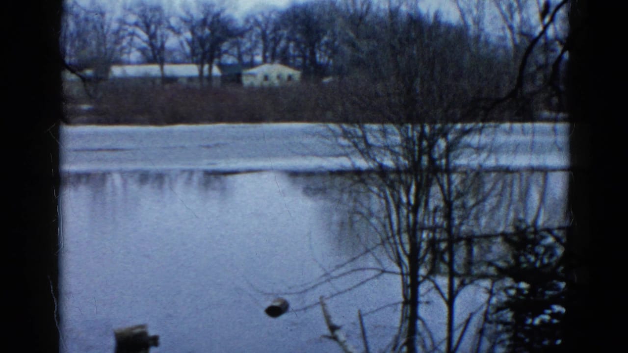 1961 Wauconda, Illinois home-movie frame of a frozen pond, bare trees, shoreline brush, and a small white house across the water.