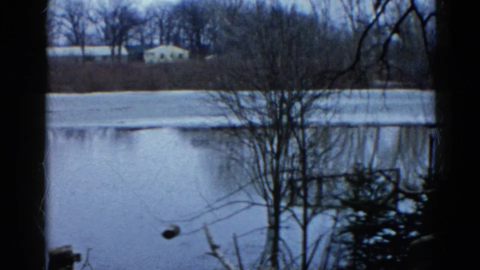 1961 Wauconda, Illinois view across a frozen pond toward bare woods and a small white house, with dark tree trunks at the frame edges.