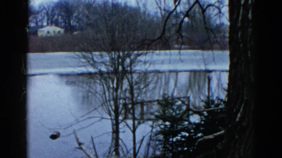 1961 Wauconda, Illinois frame showing a frozen pond through leafless branches, with shoreline brush and a small house on the far side.