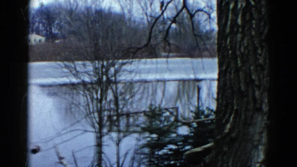 1961 Wauconda, Illinois home-movie frame of a frozen pond bordered by bare trees, dark foreground trunks, and a distant light-colored house.