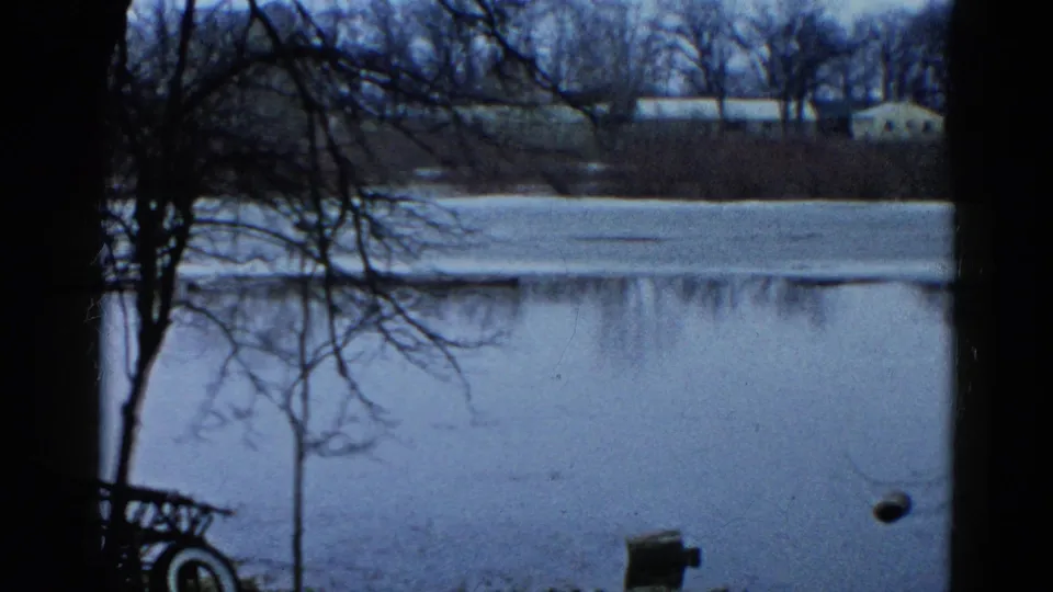 1961 Wauconda, Illinois 8mm frame of a frozen lake viewed through foreground trees, with small buildings across the shoreline.