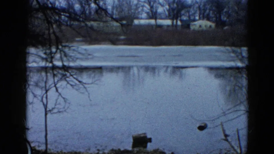 Home-movie frame from Wauconda, Illinois showing a cold lakeside view, foreground trees, ice, open water, and distant buildings.