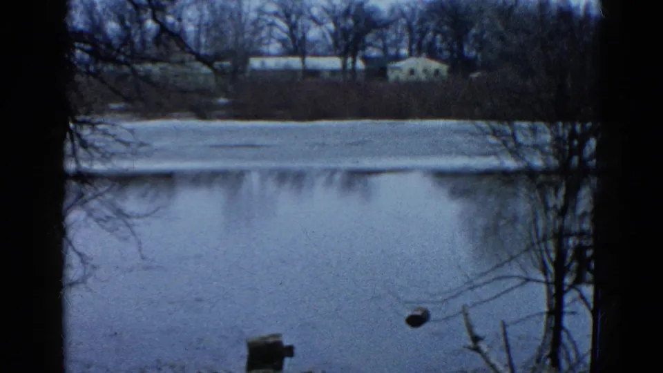 1961 8mm view of a partially frozen Wauconda lake, with a dark tree edge, distant buildings, and a small wooden foreground structure.