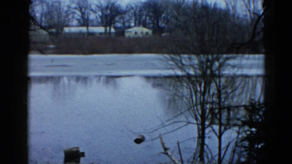Wauconda, Illinois home-movie frame showing winter trees, a frozen lake surface, open water, and distant low shoreline buildings.