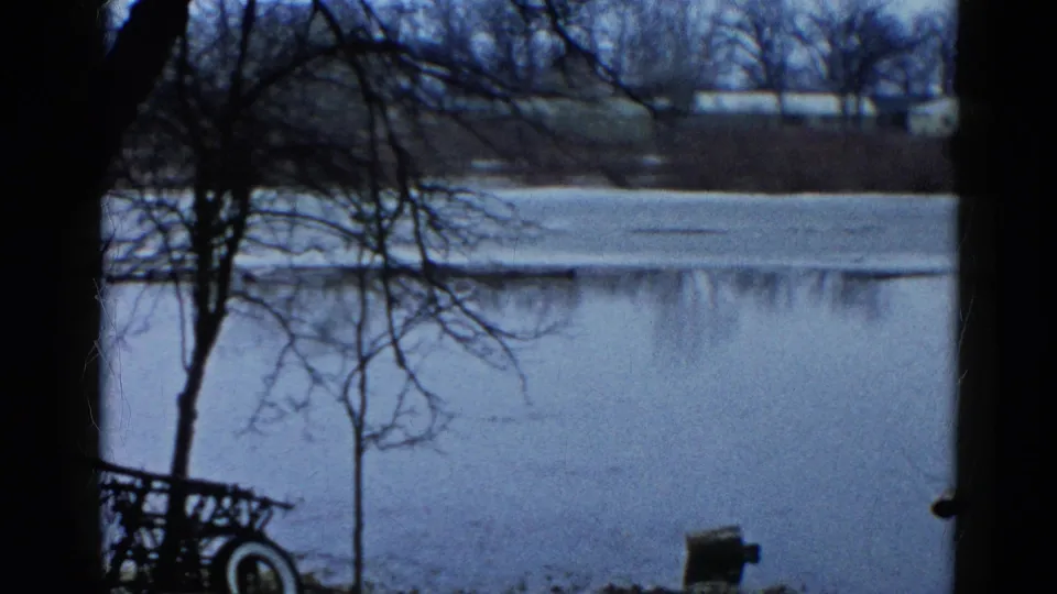 1961 Wauconda, Illinois 8mm frame with still water, a pale shoreline, dark foreground trees, and a small cart at lower left.