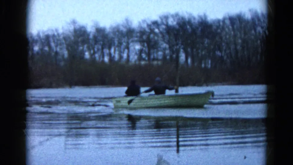 In Wauconda, Illinois, 1961 home-movie footage, two men sit in a pale rowboat on a calm lake with bare trees beyond.