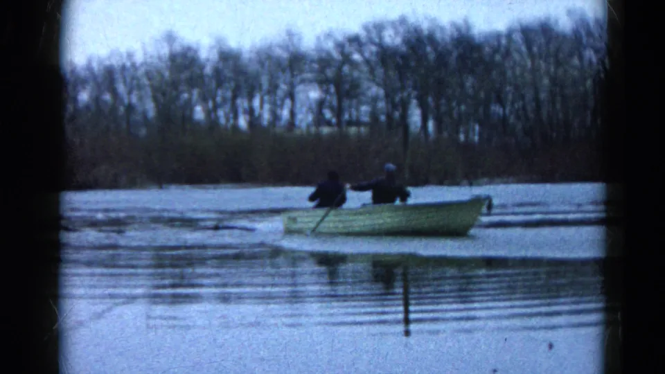 Two men in dark clothing travel in a pale rowboat across calm water in 1961 Wauconda, with a bare wooded shoreline behind them.