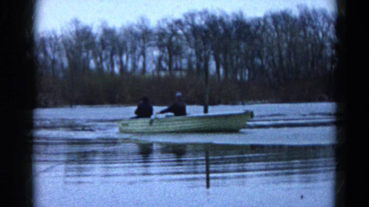Two people sit in a small rowboat on icy water with bare trees in the background.