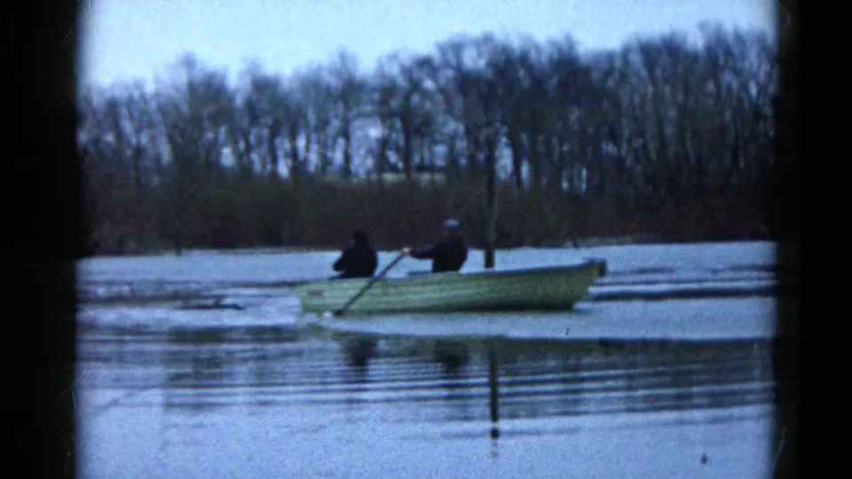 Two bundled people move across an icy Wauconda lake in a small rowboat, with one person appearing to row under an overcast sky.
