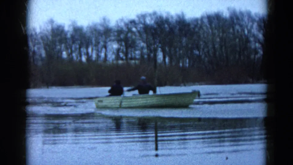 Home-movie frame from Wauconda, Illinois, showing a pale rowboat with two people crossing cold water near floating ice.