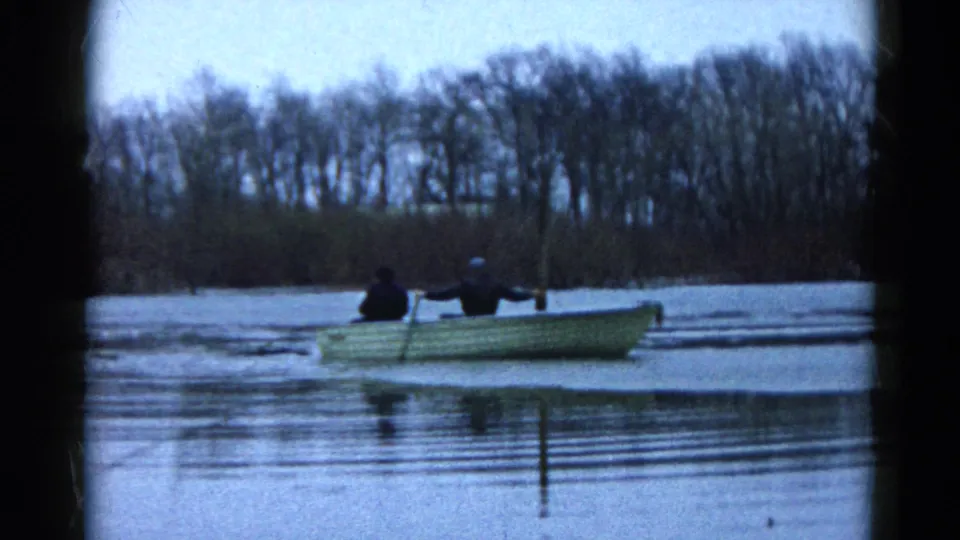 1961 8mm frame of two people in a small rowboat on an icy Wauconda lake, with ripples, bare trees, and cool winter light.