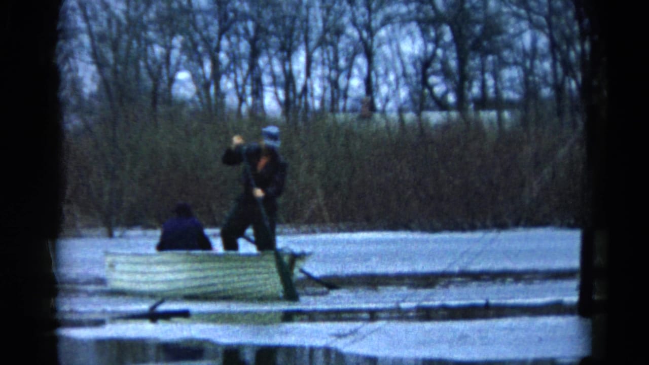 Two people in a pale rowboat on calm water near reeds and bare trees.