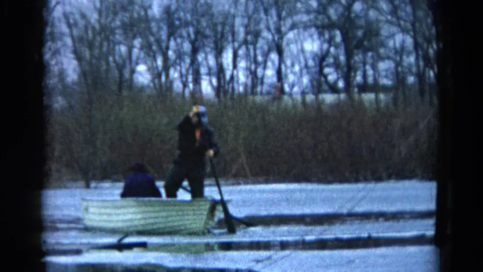 Two people remain in a small rowboat on a quiet lake, with the standing rower framed against reeds and leafless trees.