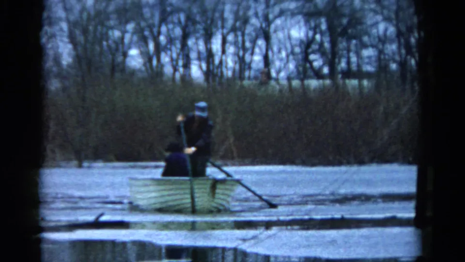 The 1961 Wauconda home-movie frame shows a man balancing in a rowboat and pushing an oar into icy pond water.