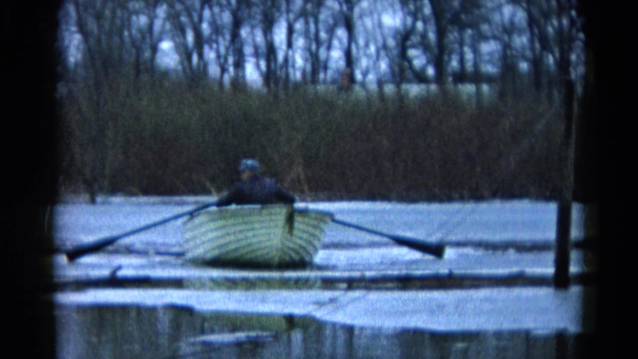 Man seated in a small rowboat on calm water with long oars extended.