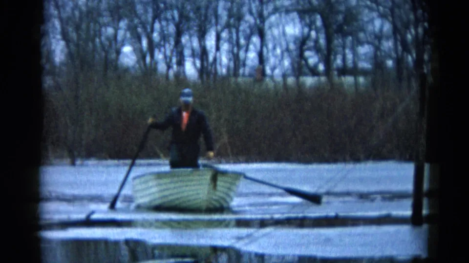A man stands in the rowboat, balancing with one oar angled down as bare trees and shoreline brush fill the background.