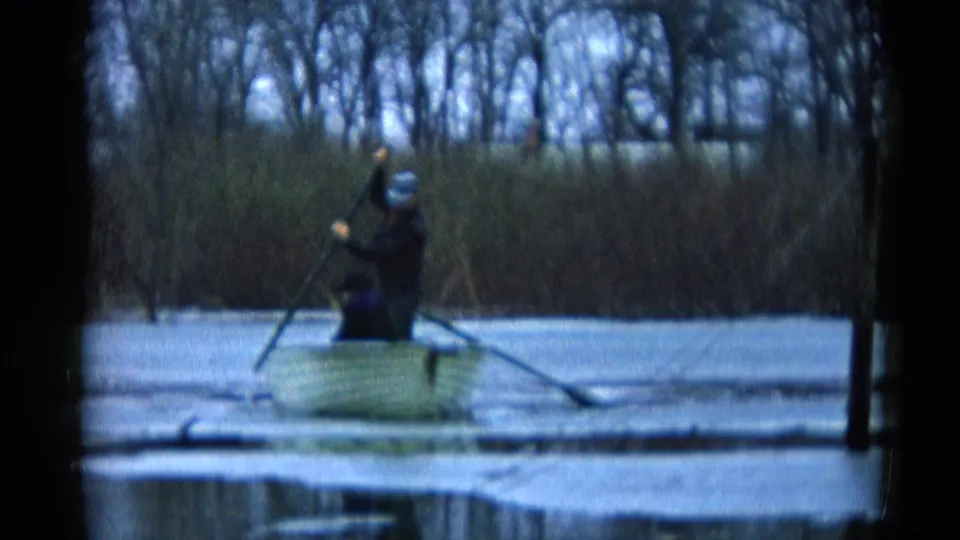The man remains standing in the small boat, lifting one oar while the other extends across the calm water near the wooded shore.